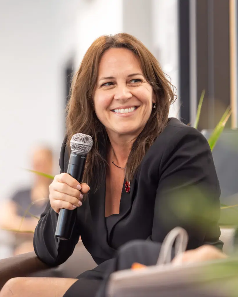 Intervenante souriante tenant un microphone lors d'une conférence. / Smiling speaker holding a microphone during a conference.