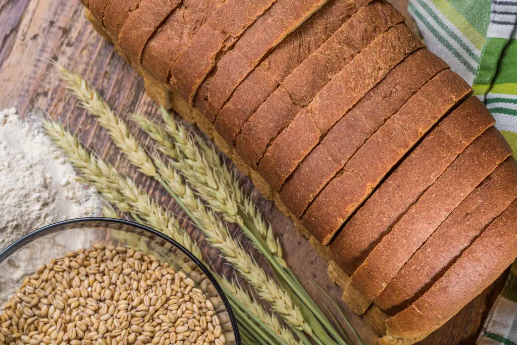 Vue en plongée d'un pain tranché, d'épis de blé, de farine et d'un bol de grains sur une table en bois. / Top-down view of sliced bread, wheat stalks, flour, and a bowl of grains on a wooden table.