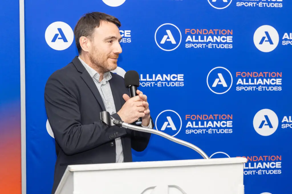 Conférencier au micro lors d’un événement de la Fondation Alliance Sport-Études devant un fond de marque. / Speaker holding a microphone at an Alliance Sport-Études Foundation event in front of a branded backdrop.