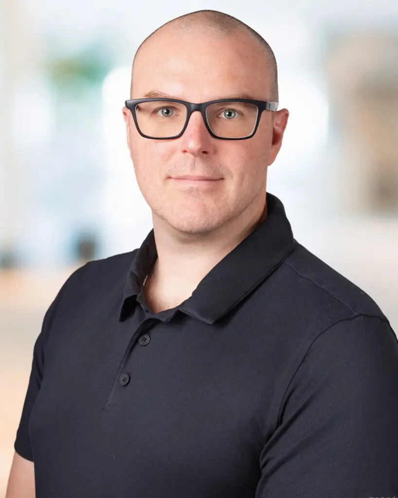 Portrait corporatif d’un homme portant des lunettes, sur fond clair et flou. / Corporate headshot of a man wearing glasses with a bright, blurred background.