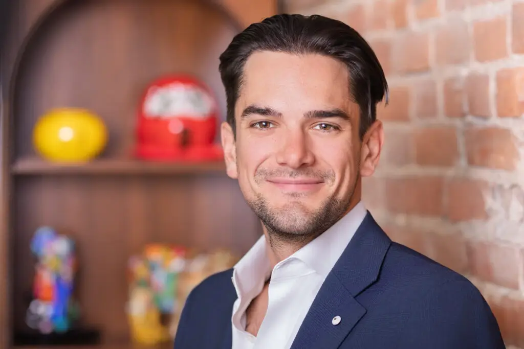 Portrait corporatif d’un homme en veston bleu sur fond de mur de brique, éclairage doux et arrière-plan flou. / Corporate portrait of a man in a blue suit against a brick wall background with soft lighting and blurred background.